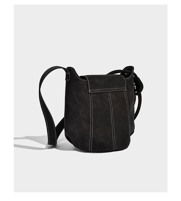 Small matte textured bucket crossbody bag with flap closure, gold-tone bar hardware, contrast stitching, and pompom tassel