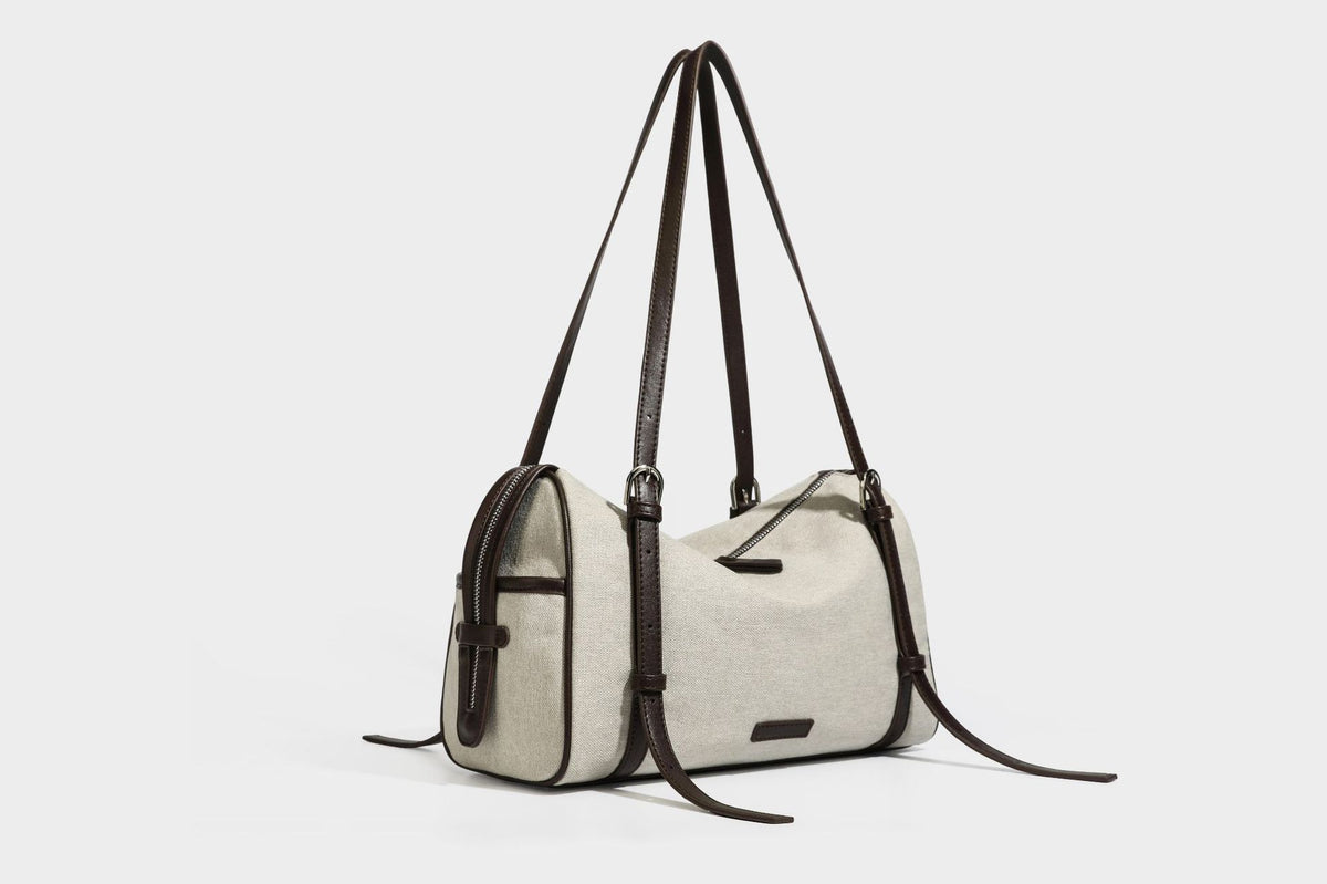Beige canvas barrel bag with dark brown leather-look trim, silver-tone hardware, top zip closure, and adjustable shoulder