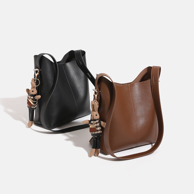 Smooth leather-look bucket bag with long flat shoulder strap, open top, gold-tone hardware, and decorative rabbit charm