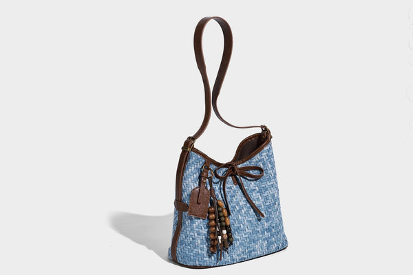 Light blue woven bucket bag with brown leather-look trim, gold-tone hardware, adjustable shoulder strap, and decorative bead