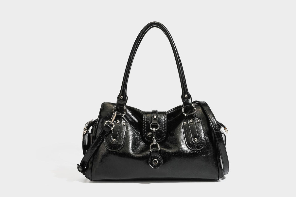 Glänzende, geknautschte Barrel-Satchel mit zwei oberen Tragegriffen, vorderem Klappen-Riemen-Panel, Ring- und Nietenbeschlägen sowie einem abnehmbaren