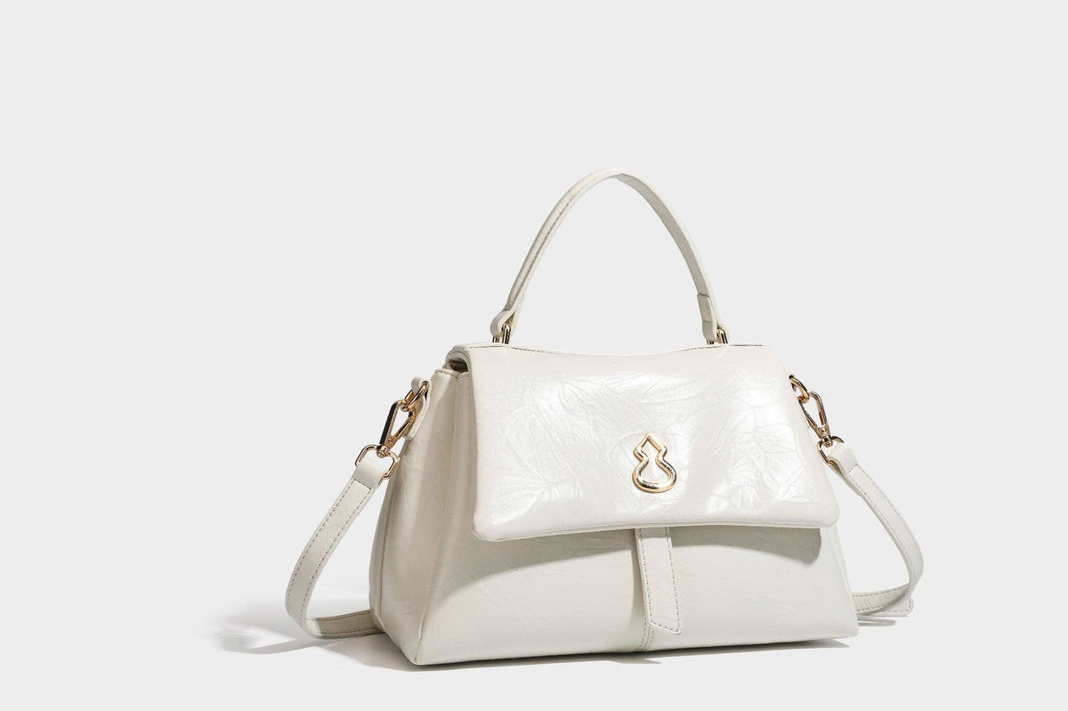Structured black satchel bag with glossy crinkled leather-look finish, gold-tone emblem flap closure, slim top handle, and