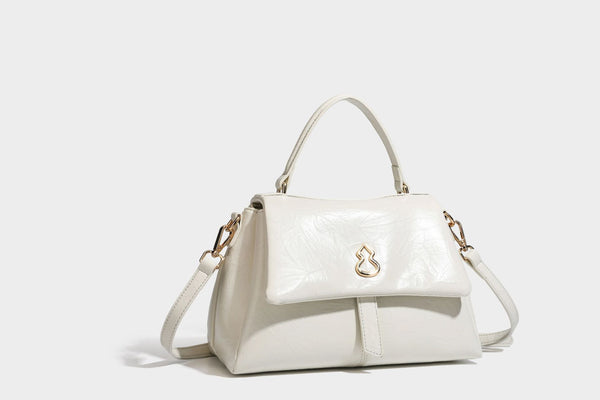Structured black satchel bag with glossy crinkled leather-look finish, gold-tone emblem flap closure, slim top handle, and