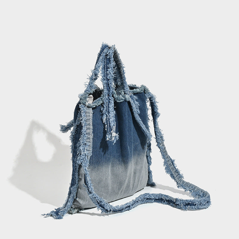 Washed denim fringe crossbody bag with indigo-to-pale-blue gradient and raw fringe trim along edges and shoulder strap