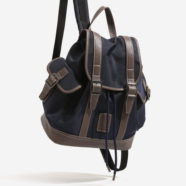 Deep navy canvas backpack with brown textured leather-look trim, drawstring flap closure, buckle-strap front detail, and