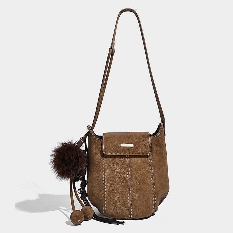 Small matte textured bucket crossbody bag with flap closure, gold-tone bar hardware, contrast stitching, and pompom tassel