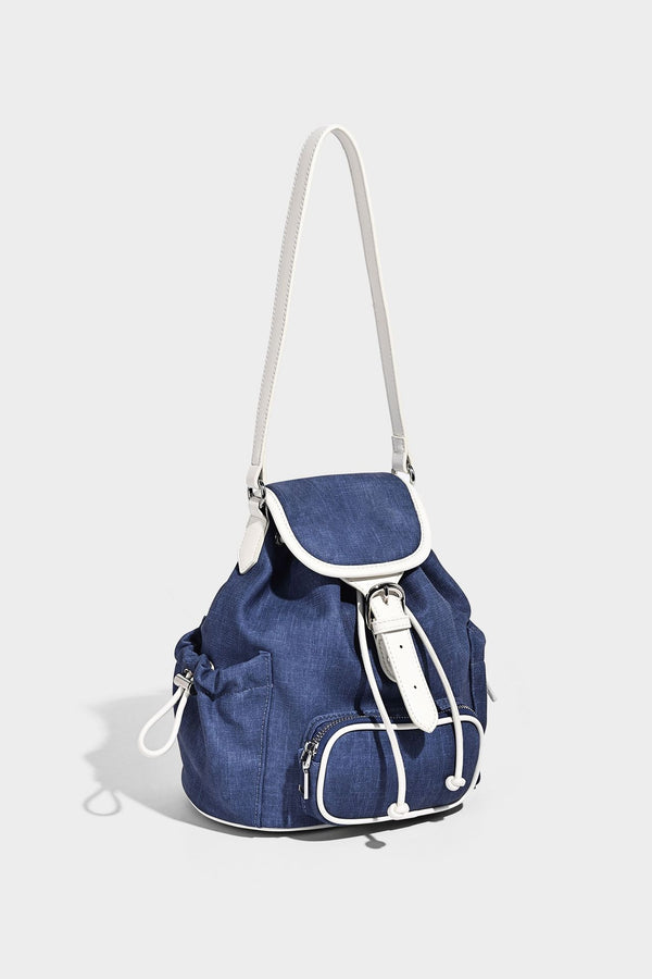 Compact denim drawstring backpack with white leather-look piping, flap-top buckle closure, front zip pocket, and silver-tone