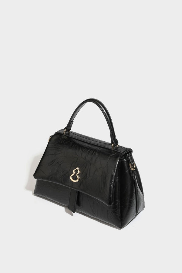 Structured black satchel bag with glossy crinkled leather-look finish, gold-tone emblem flap closure, slim top handle, and