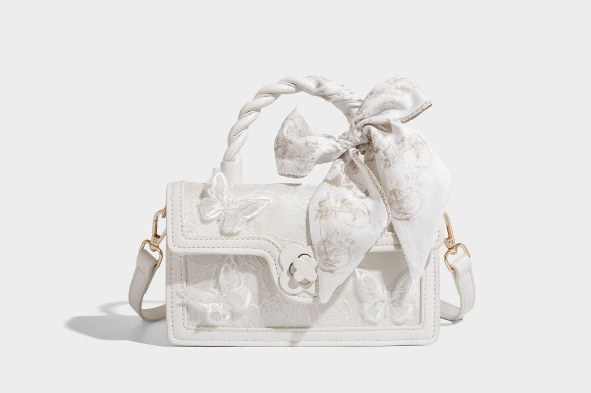 Small structured white flap bag with butterfly embroidery, flower-shaped turn-lock clasp, braided top handle with scarf bow