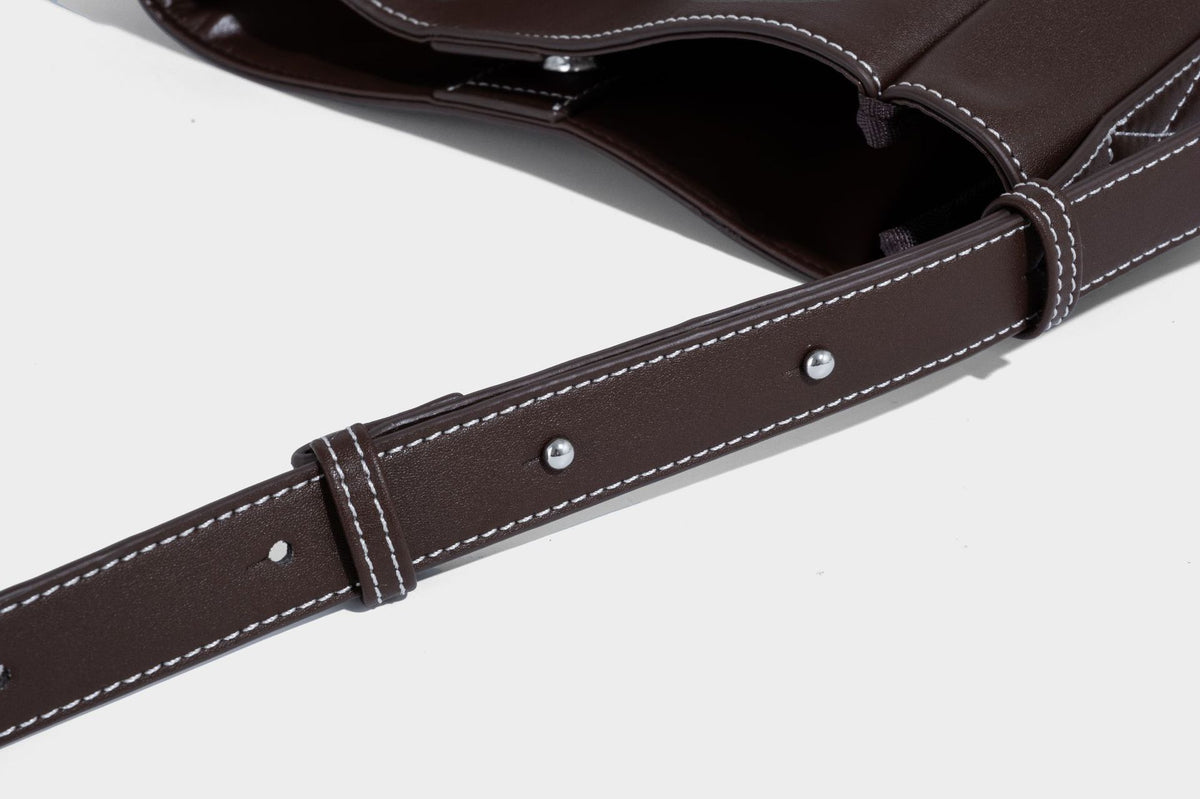 Structured dark brown leather-look bucket bag with white contrast stitching, front wraparound belt buckle strap, and long