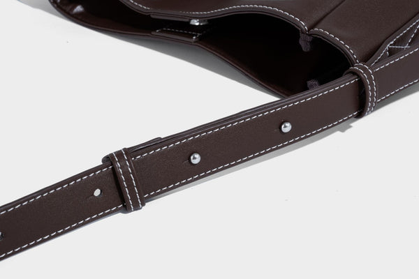 Structured dark brown leather-look bucket bag with white contrast stitching, front wraparound belt buckle strap, and long