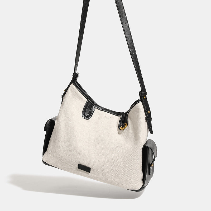 Off-white canvas shoulder bag with black leather-look trim, gold-tone ring hardware, top zip closure, side flap pocket, and