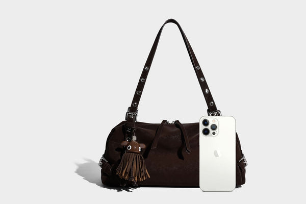 Weiche, zylinderförmige Schultertasche in Dunkelbraun mit silberfarbenen Riemen mit Ösen, Reißverschluss oben, seitlichen Schnallenbeschlägen und Quaste