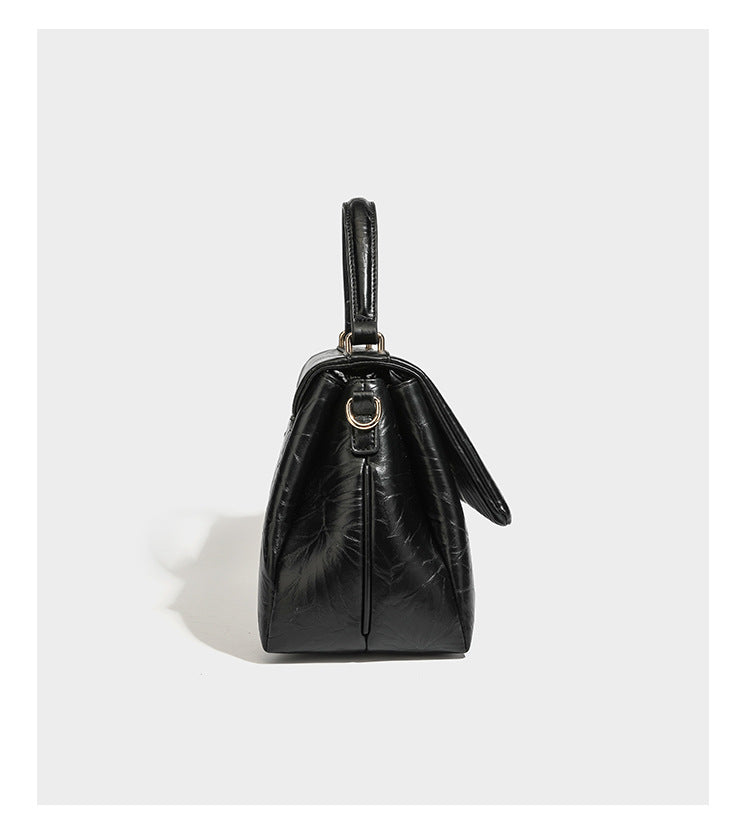 Structured black satchel bag with glossy crinkled leather-look finish, gold-tone emblem flap closure, slim top handle, and