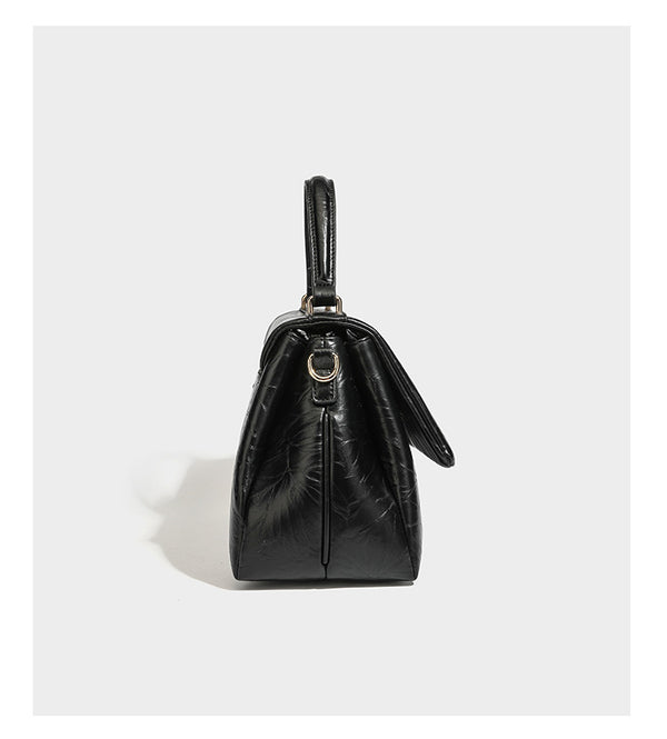 Structured black satchel bag with glossy crinkled leather-look finish, gold-tone emblem flap closure, slim top handle, and