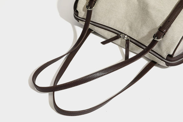Beige canvas barrel bag with dark brown leather-look trim, silver-tone hardware, top zip closure, and adjustable shoulder