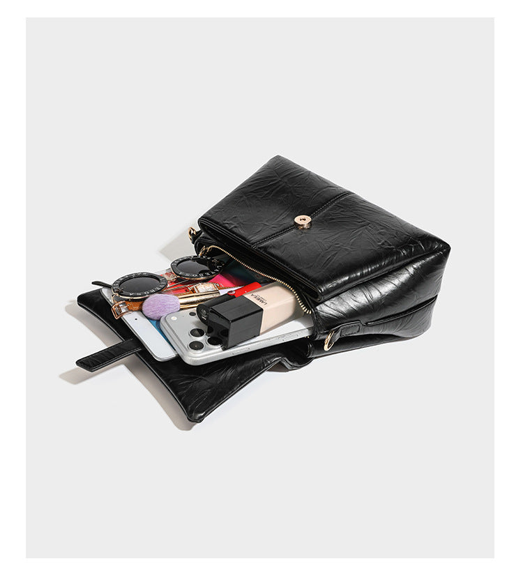 Structured black satchel bag with glossy crinkled leather-look finish, gold-tone emblem flap closure, slim top handle, and