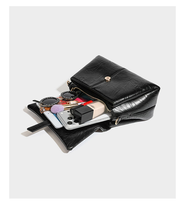 Structured black satchel bag with glossy crinkled leather-look finish, gold-tone emblem flap closure, slim top handle, and