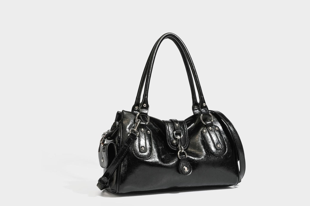 Glänzende, geknautschte Barrel-Satchel mit zwei oberen Tragegriffen, vorderem Klappen-Riemen-Panel, Ring- und Nietenbeschlägen sowie einem abnehmbaren