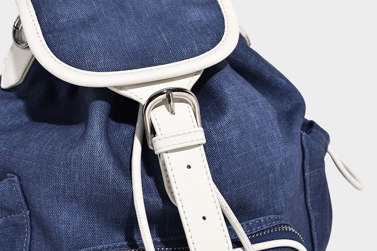 Compact denim drawstring backpack with white leather-look piping, flap-top buckle closure, front zip pocket, and silver-tone