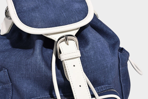 Compact denim drawstring backpack with white leather-look piping, flap-top buckle closure, front zip pocket, and silver-tone