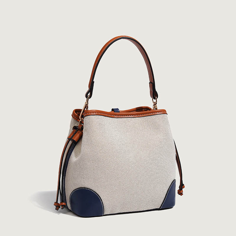 Cream woven-texture canvas bucket bag with navy accent panels, tan leather-look trim, gold-tone chain strap, and tassel
