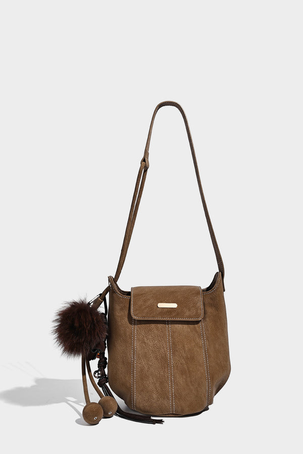 Small matte textured bucket crossbody bag with flap closure, gold-tone bar hardware, contrast stitching, and pompom tassel
