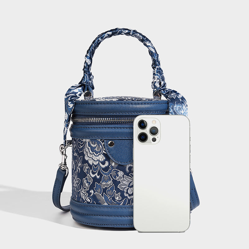 Small cylindrical barrel bag in blue-and-white floral jacquard with leather-look trim, scarf-wrapped top handle, silver-tone