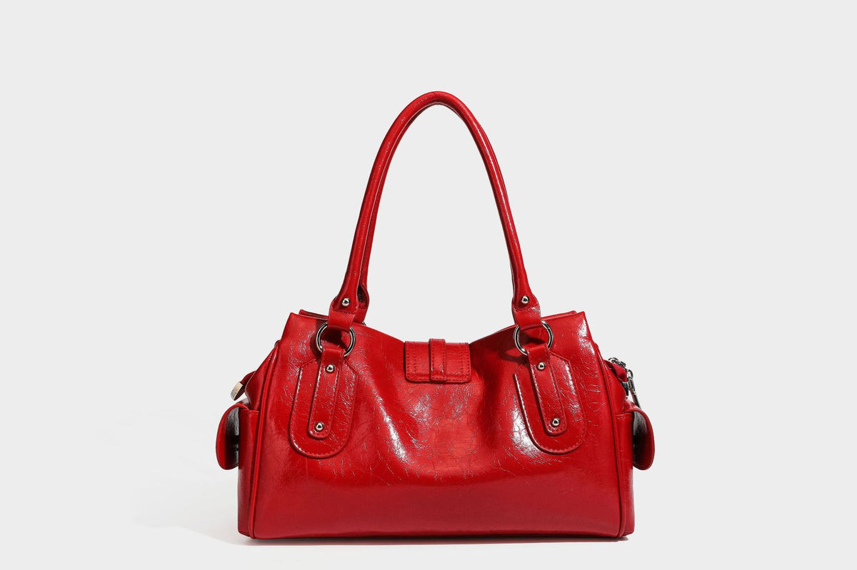 Glänzende, geknautschte Barrel-Satchel mit zwei oberen Tragegriffen, vorderem Klappen-Riemen-Panel, Ring- und Nietenbeschlägen sowie einem abnehmbaren