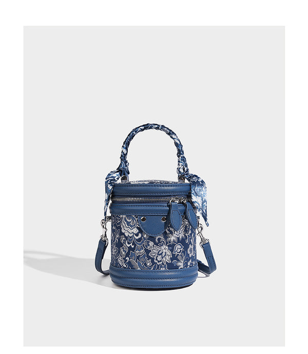 Small cylindrical barrel bag in blue-and-white floral jacquard with leather-look trim, scarf-wrapped top handle, silver-tone