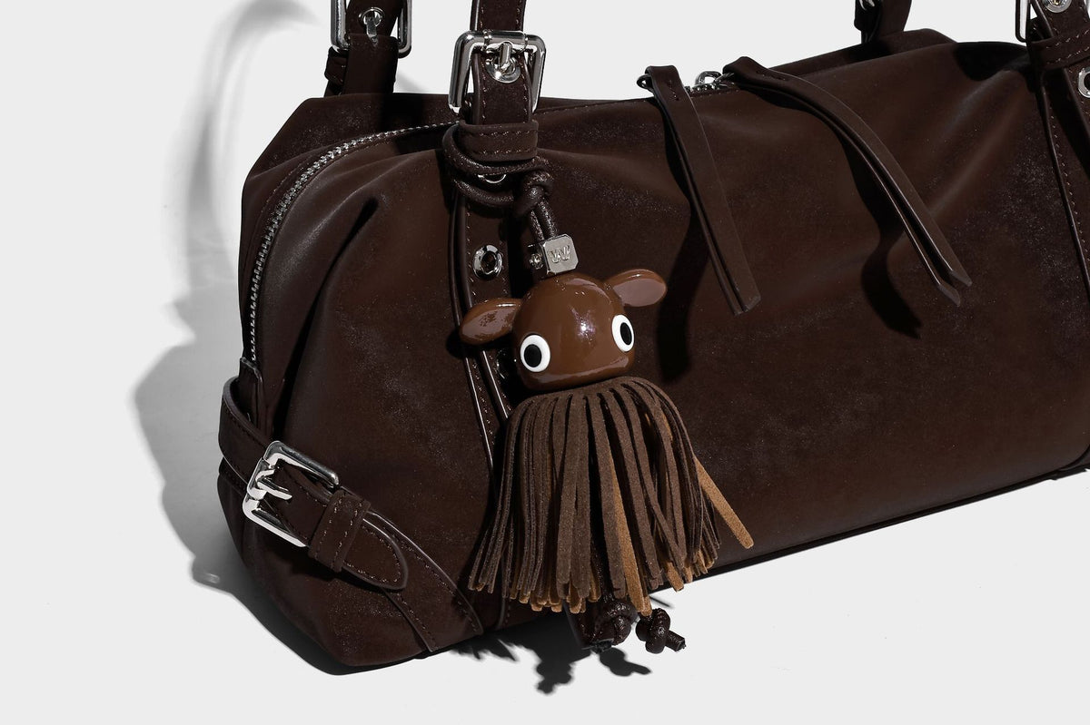 Weiche, zylinderförmige Schultertasche in Dunkelbraun mit silberfarbenen Riemen mit Ösen, Reißverschluss oben, seitlichen Schnallenbeschlägen und Quaste