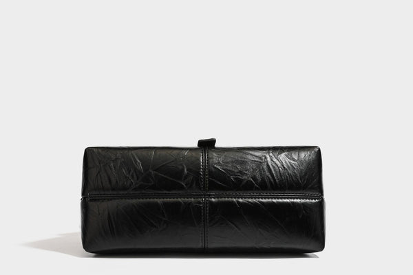 Structured black satchel bag with glossy crinkled leather-look finish, gold-tone emblem flap closure, slim top handle, and
