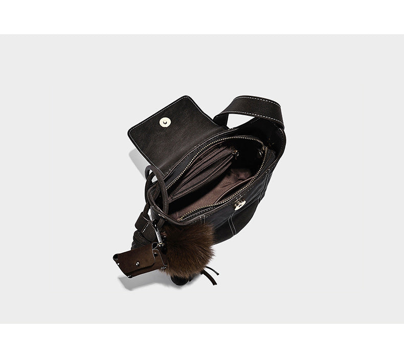Small matte textured bucket crossbody bag with flap closure, gold-tone bar hardware, contrast stitching, and pompom tassel