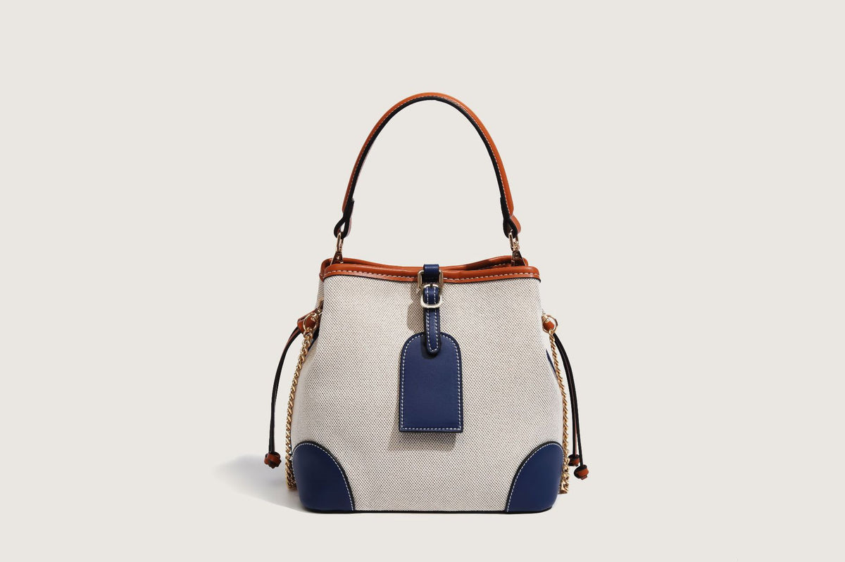 Cream woven-texture canvas bucket bag with navy accent panels, tan leather-look trim, gold-tone chain strap, and tassel