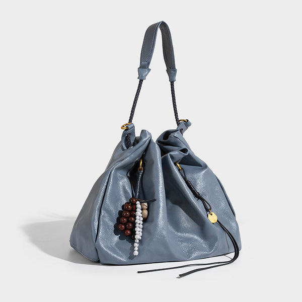 Cream slouchy drawstring bucket bag with braided cord, beaded tassel charm, and gold-tone ring hardware, front view