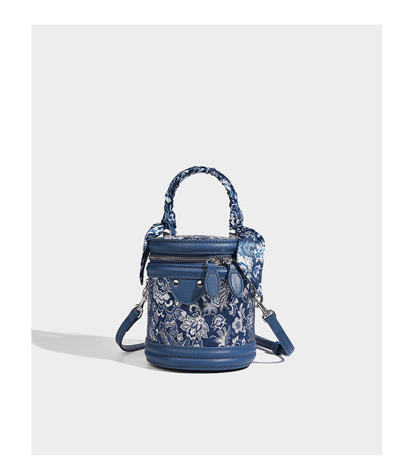 Small cylindrical barrel bag in blue-and-white floral jacquard with leather-look trim, scarf-wrapped top handle, silver-tone