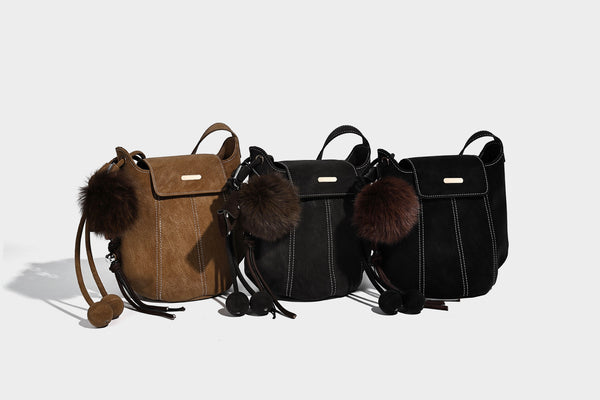 Small matte textured bucket crossbody bag with flap closure, gold-tone bar hardware, contrast stitching, and pompom tassel