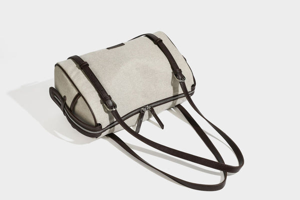 Beige canvas barrel bag with dark brown leather-look trim, silver-tone hardware, top zip closure, and adjustable shoulder