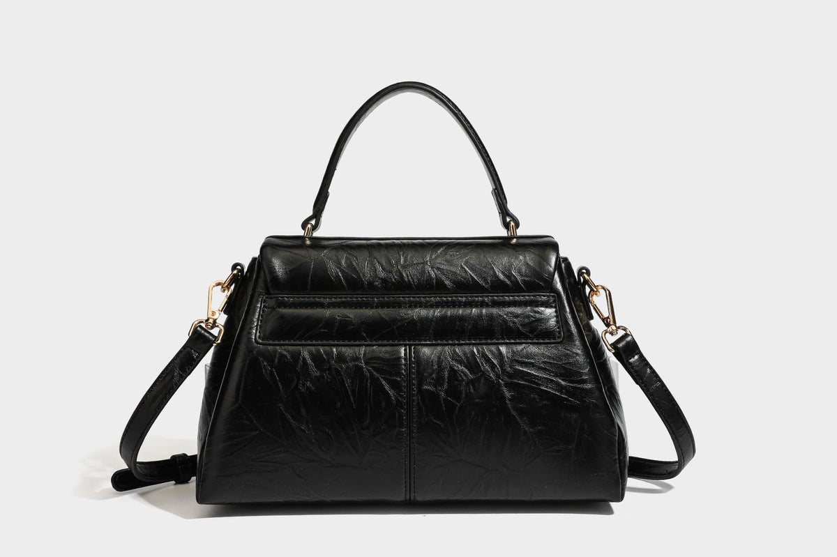 Structured black satchel bag with glossy crinkled leather-look finish, gold-tone emblem flap closure, slim top handle, and