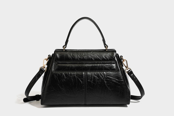 Structured black satchel bag with glossy crinkled leather-look finish, gold-tone emblem flap closure, slim top handle, and
