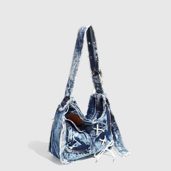 Slouchy distressed denim shoulder bag with frayed edges, white lace-up cross detail, front snap pocket, and long shoulder