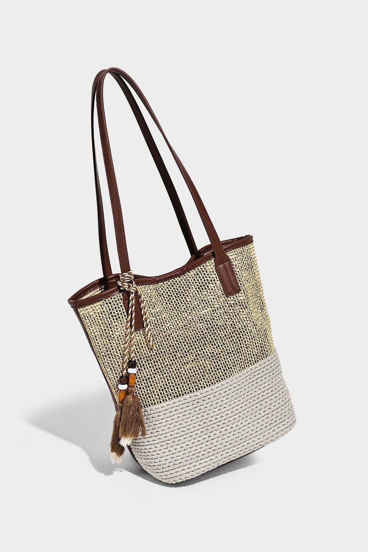 Woven straw-look tote bag in tan and cream with dark brown leather-look trim, long flat shoulder straps, and tassel bead