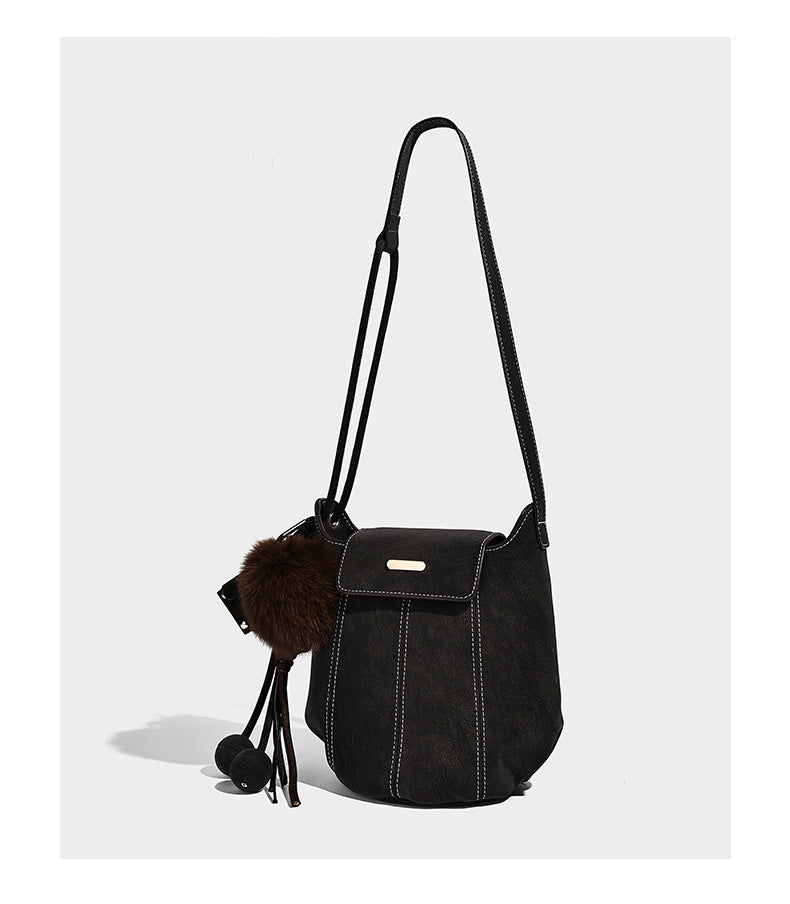 Small matte textured bucket crossbody bag with flap closure, gold-tone bar hardware, contrast stitching, and pompom tassel