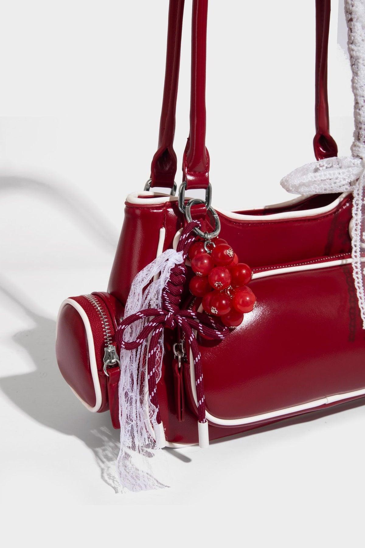 Kleine, formstabile, glänzende Schultertasche mit weißer Paspelierung, Spitzenschleife und Anhänger mit roter Perle, seitlichen Reißverschlusstaschen und silberfarbenem