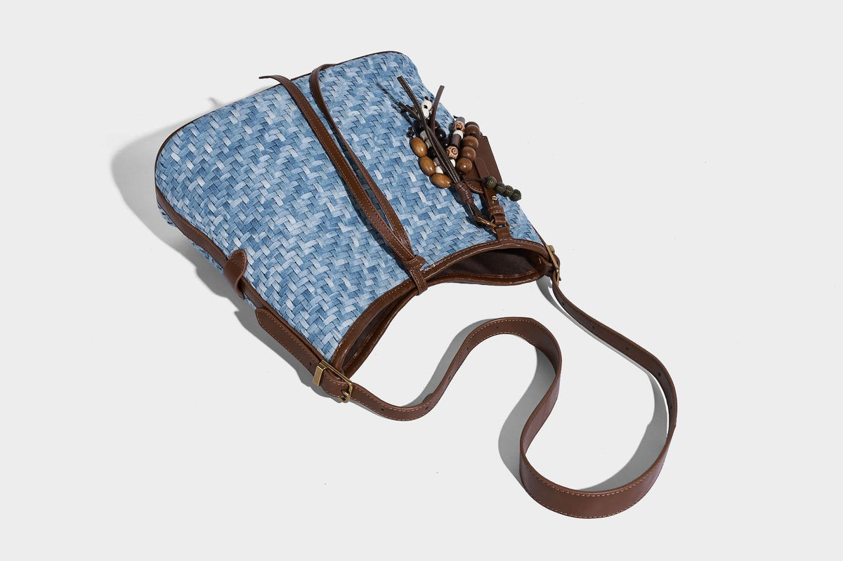 Light blue woven bucket bag with brown leather-look trim, gold-tone hardware, adjustable shoulder strap, and decorative bead