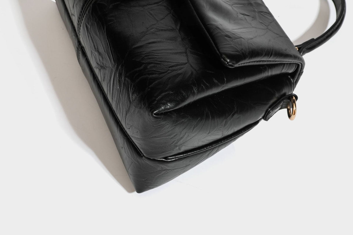 Structured black satchel bag with glossy crinkled leather-look finish, gold-tone emblem flap closure, slim top handle, and