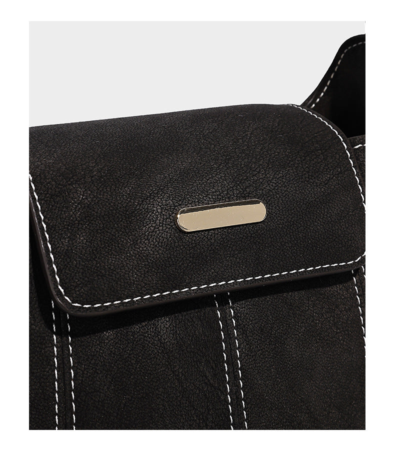 Small matte textured bucket crossbody bag with flap closure, gold-tone bar hardware, contrast stitching, and pompom tassel