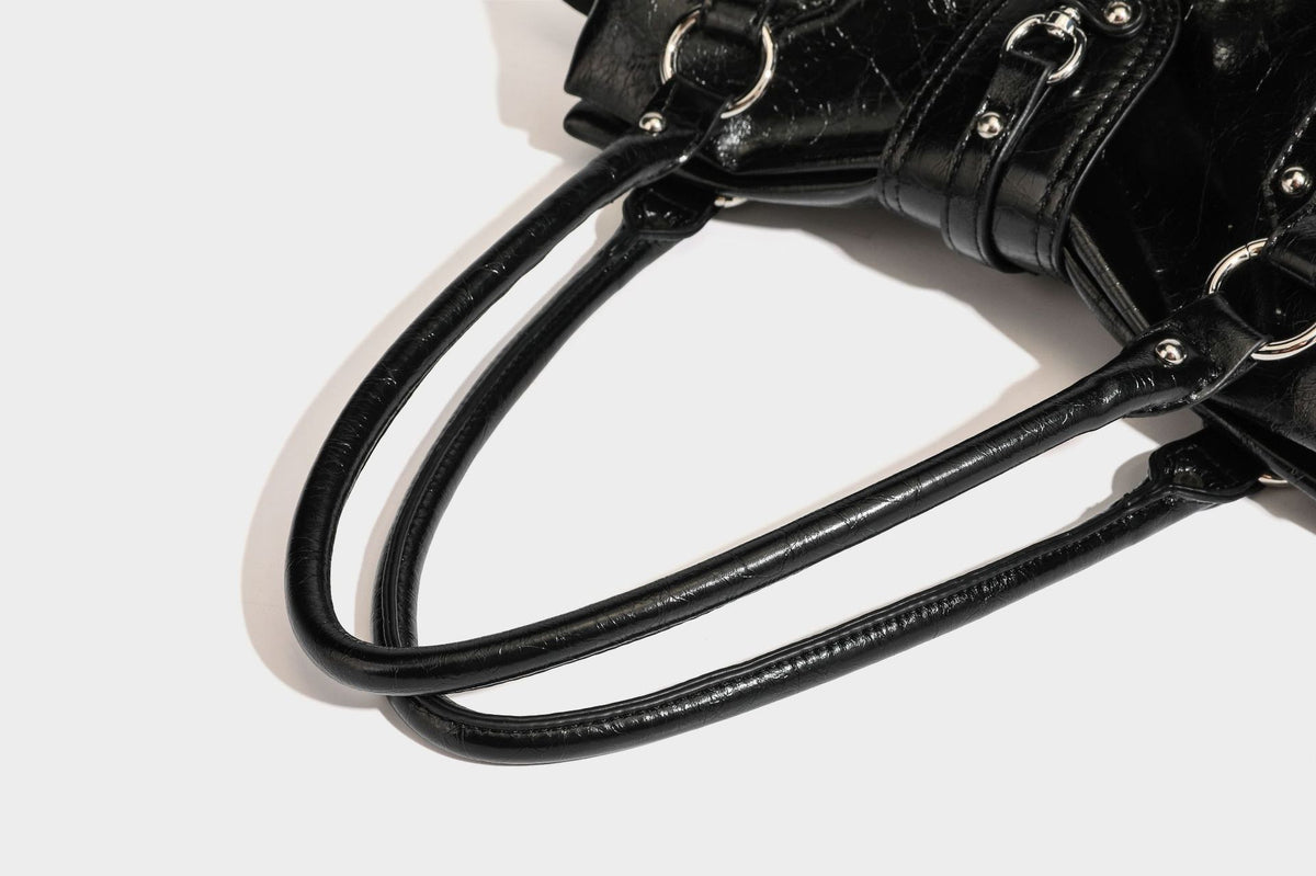 Glänzende, geknautschte Barrel-Satchel mit zwei oberen Tragegriffen, vorderem Klappen-Riemen-Panel, Ring- und Nietenbeschlägen sowie einem abnehmbaren