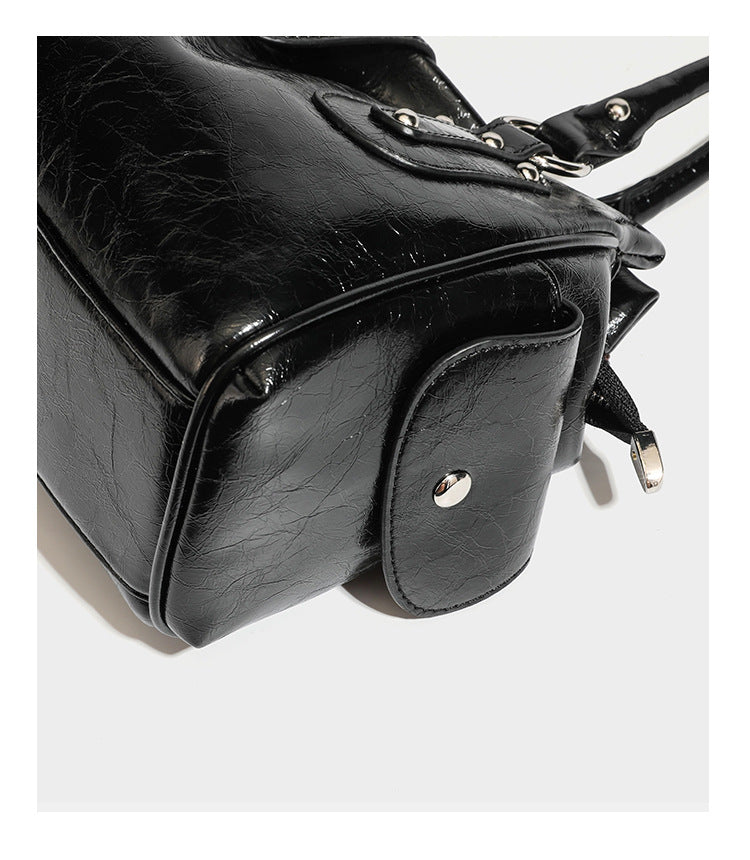 Glänzende, geknautschte Barrel-Satchel mit zwei oberen Tragegriffen, vorderem Klappen-Riemen-Panel, Ring- und Nietenbeschlägen sowie einem abnehmbaren