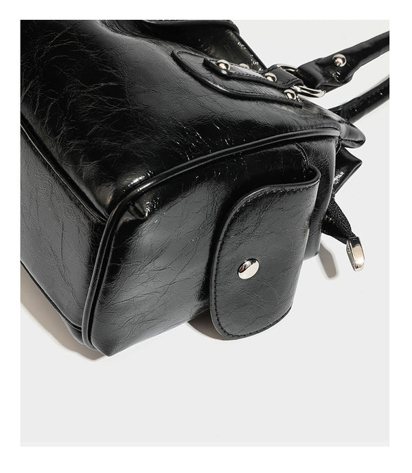 Glänzende, geknautschte Barrel-Satchel mit zwei oberen Tragegriffen, vorderem Klappen-Riemen-Panel, Ring- und Nietenbeschlägen sowie einem abnehmbaren